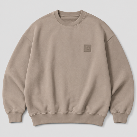 A taupe crewneck sweatshirt with a small tonal square patch on the left chest, displayed flat against a light gray background.